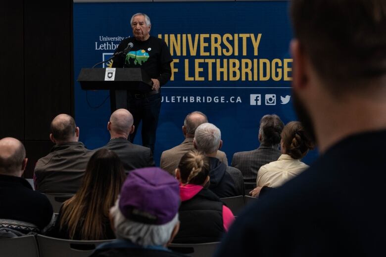 An elderly man stands on a podium addressing a crowd with a banner reading University of Lethbridge behind him. 