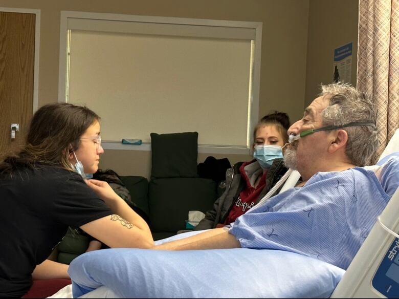 A young woman sits on a hospital bed, talking to an older man in a blue hospital gown.