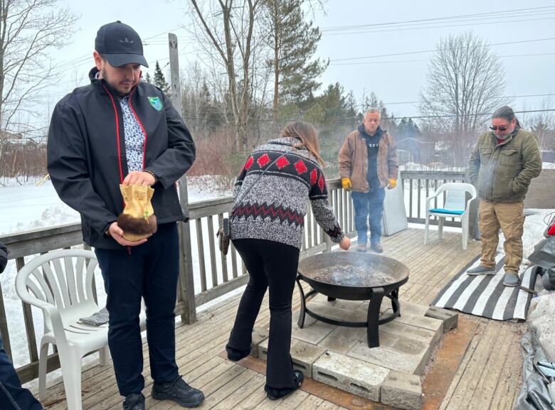 Four people stand around a fire. One woman is bending over, offering tobacco to the fire.