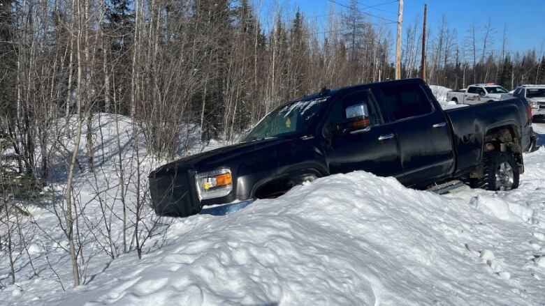 A black pickup truck is seen stuck in a snow bank along a wooded area with three RCMP vehicles behind it.