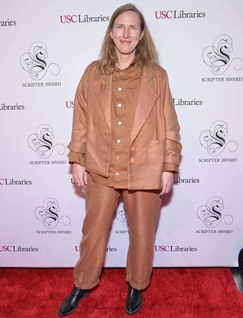 A woman with long auburn hair stands on red carpet in front of white backdrop. She wears a monochrome tan suit with black shoes. 