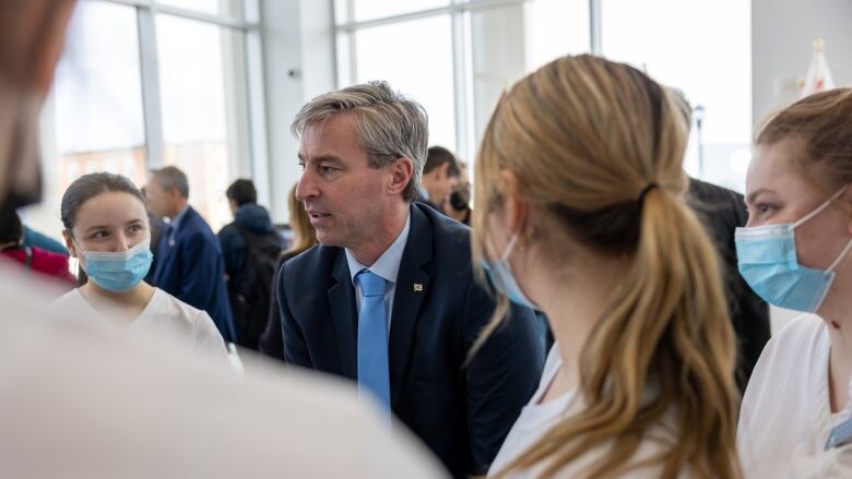 A man in a suit speaks to a group of young woman who are wearing medical face masks.