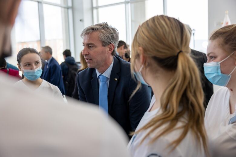 A man in a suit speaks to a group of young woman who are wearing medical face masks.