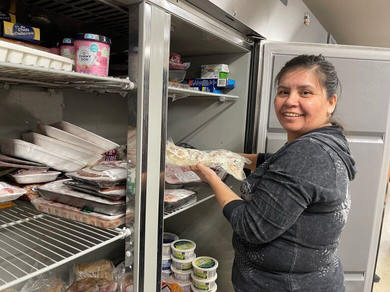 A woman smiles at the camera while taking some food out of an industrial fridge.