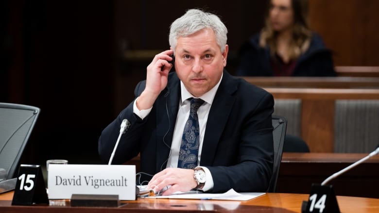 Director of the Canadian Security Intelligence Service David Vigneault sits at a desk wearing a dark suit. He is a middle-aged white man and has a focused look on his face while adjusting an ear piece that acts as a translation aid. 