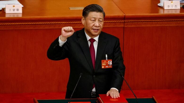 A man stands at a podium holding up a closed hand as his other hand rests on a book with a red cover.