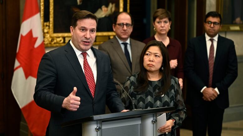 White man in suit and tie speaks from a podium flanked by an Asian woman and three other people behind them.