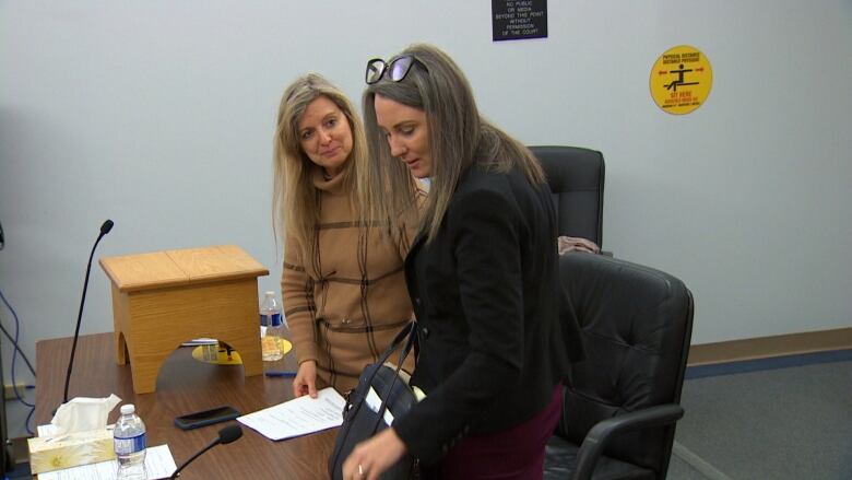 Two women standing at a table in a courtroom. The woman in the background is looking at the other with a slight smile, while the woman in the foreground is reaching for something on the table.