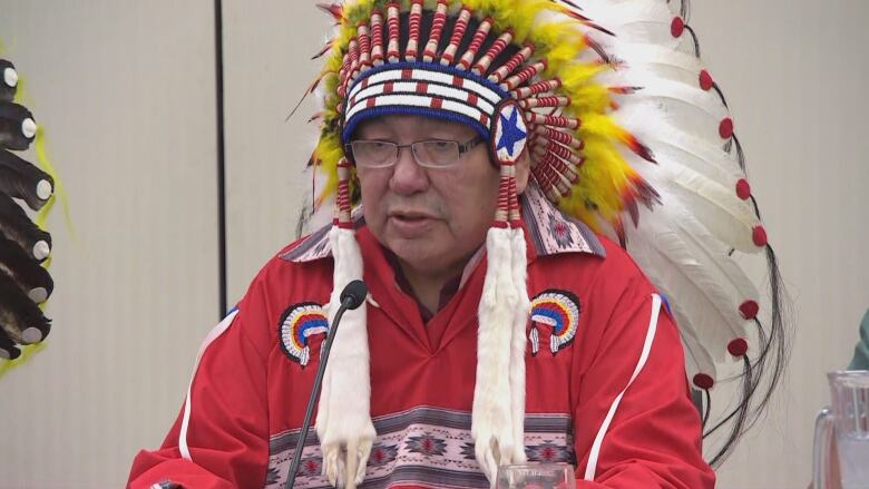 A man with glasses and a colourful First Nations headdress speaks into a microphone while sitting at a table.