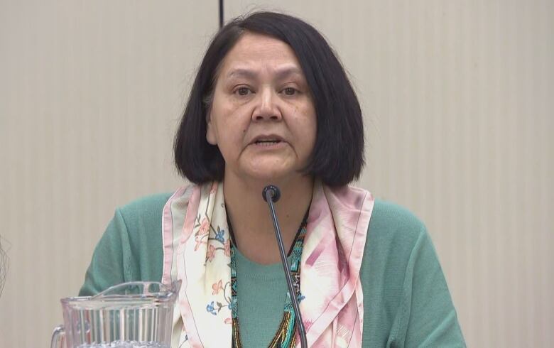 A woman with shoulder-length black hair speaks into a microphone while sitting at a table. She is wearing a green sweater and a floral-patterned scarf.