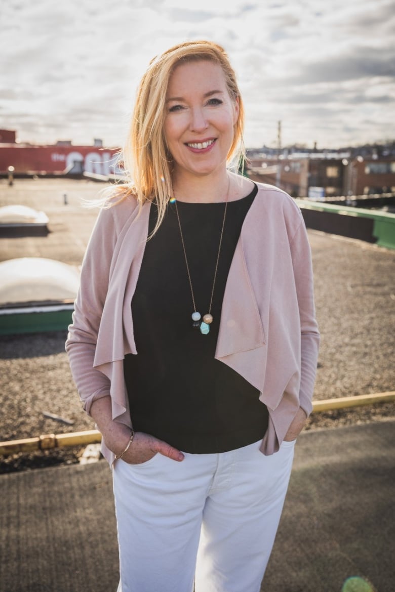 A woman with a black shirt and pink cardigan stands, looking at the camera, on a city rooftop.