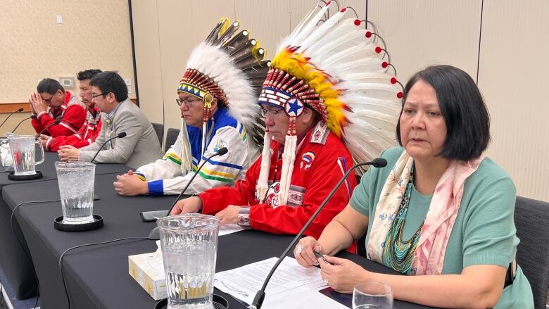 Six people sit at a long table. A woman with black hair sits at the far right of the photo and speaks into a microphone. Beside her are two men wearing First Nations headdresses.