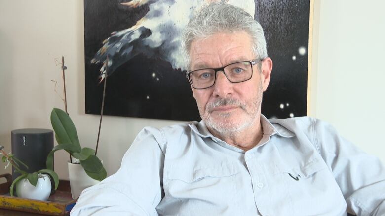 Man sits in his home with a painting and a plant behind him. 