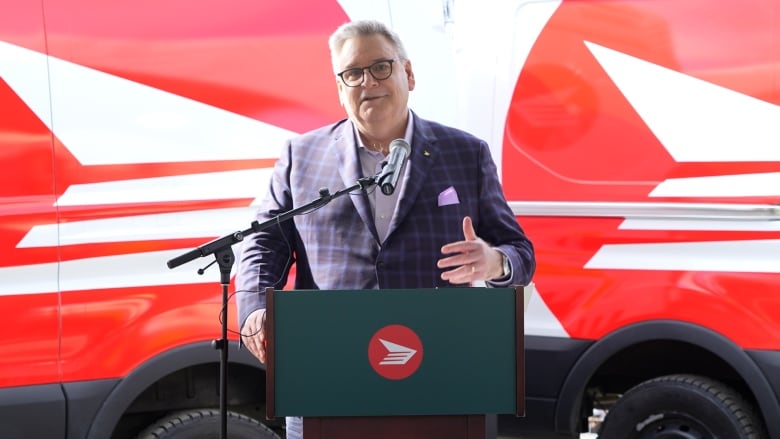 Canada Post CEO speaks at a podium with the Canada Post logo and two trucks parked in the back 