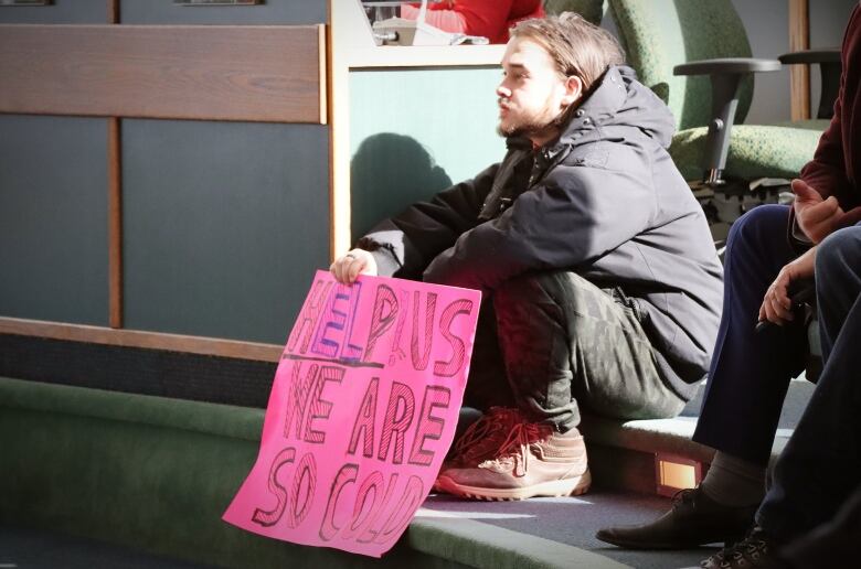 A man sits in the sun showing a sign that says help us we are so cold.