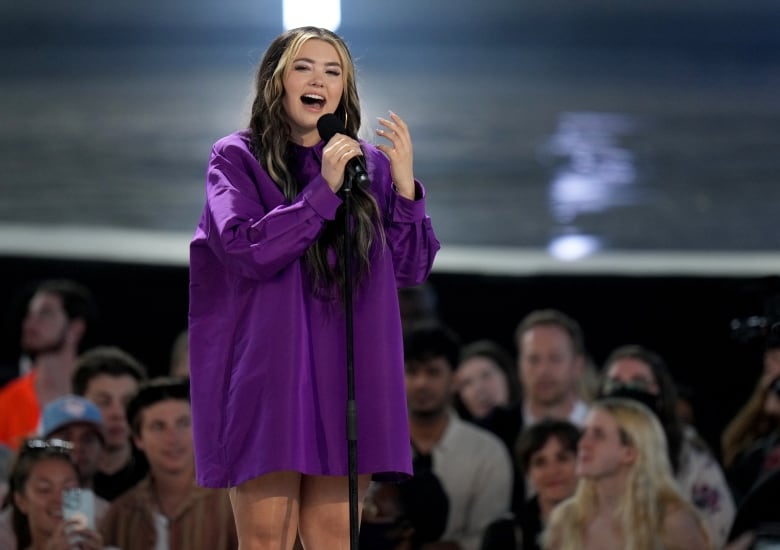 A young woman in a purple dress sings into a microphone.