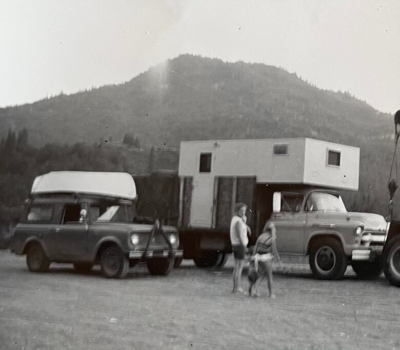 A black and white photo of a camper truck and another vehicle parked with a woman and a girl standing nearby.