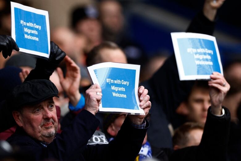 Three people hold signs above their heads in front of a crowd of seated people in a stadium. 