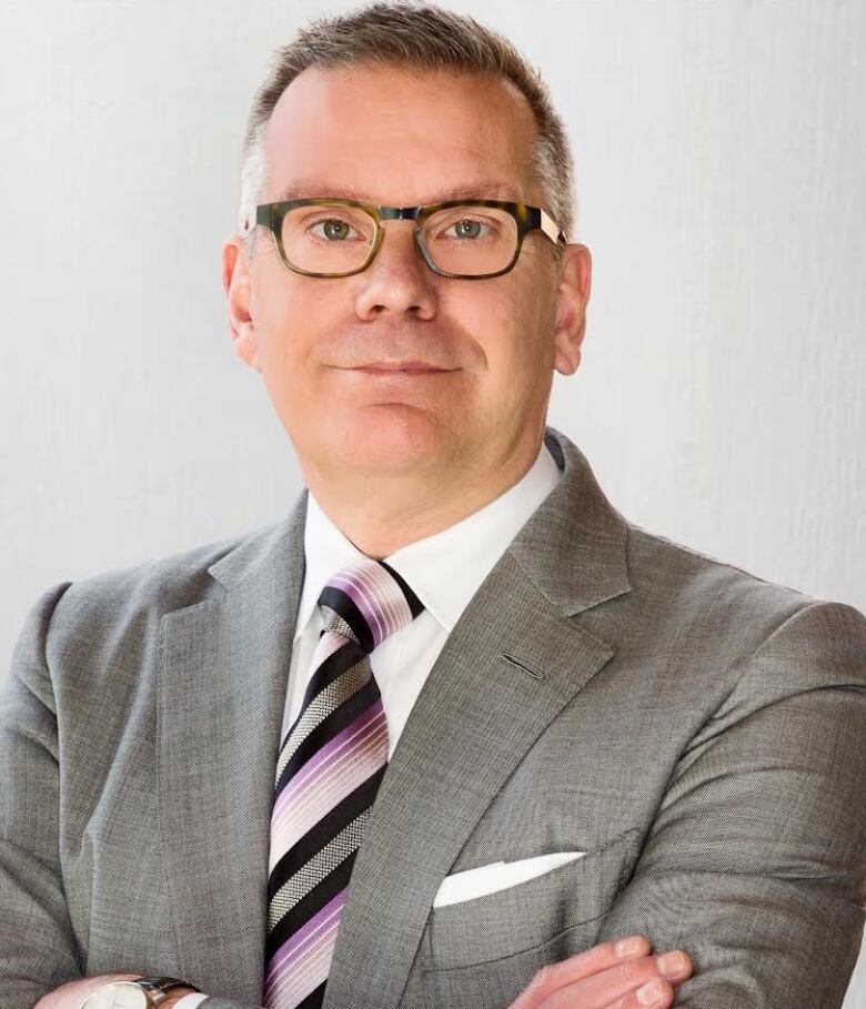 A headshot of a man wearing a grey suit. he is wearing a purple and black tie. he has grey hair and is wearing glasses