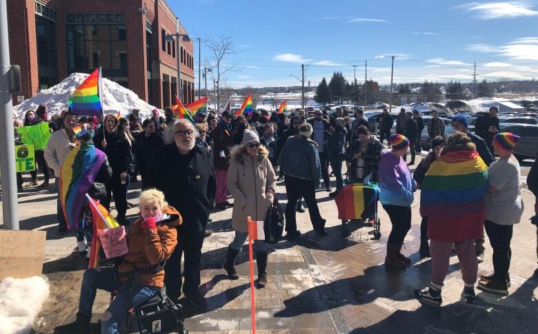 A crowd of people is shown on the sidewalk and nearby street outside a brick building. Many are carrying rainbow LGBTQ flags or wearing rainbow hats. A few towards the left of the photo are holding green poster board signs.