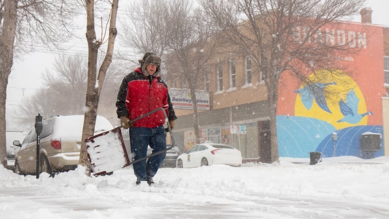 A man is seen with a snow shovel, walking down a snowy sidewalk.