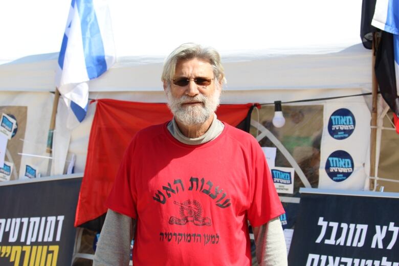 Man with grey hair and beard wearing sunglasses and red T-shirt stands outside a protest booth