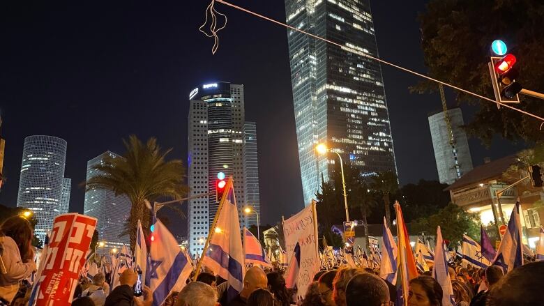 Protesters hold Israeli flags and signs at night on the street under office towers with lights.