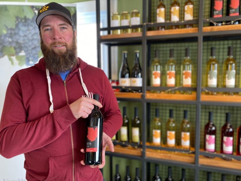 A man with a red sweatshirt stands in front of a shelf of wines, holding a bottle of red wine.