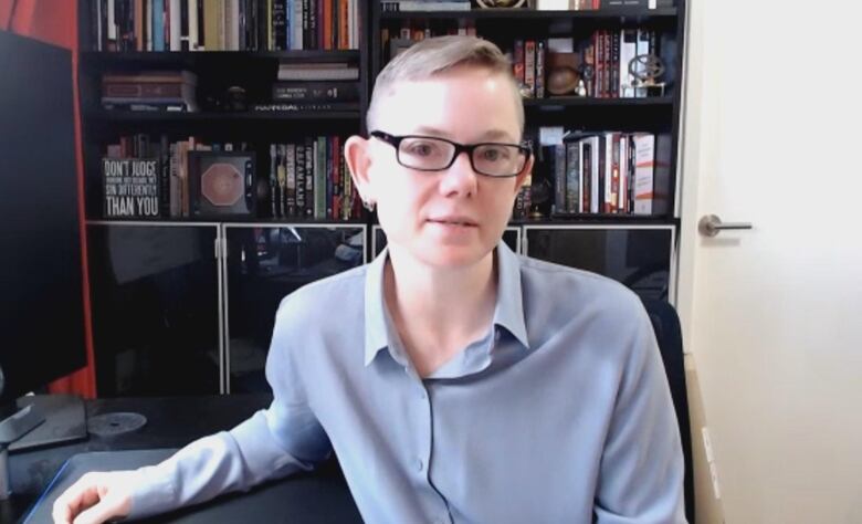 A person sits in an office with books behind them. they are wearing a blue button up shirt 