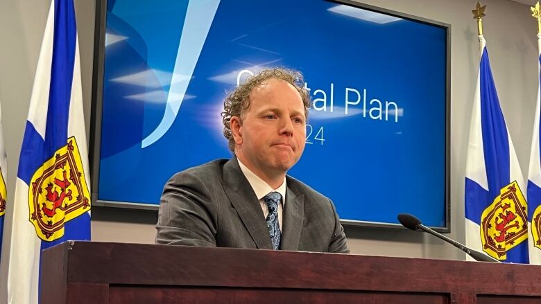 A man wearing a grey blazer with a white shirt and blue tie sits behind a desk between two Nova Scotia flags.