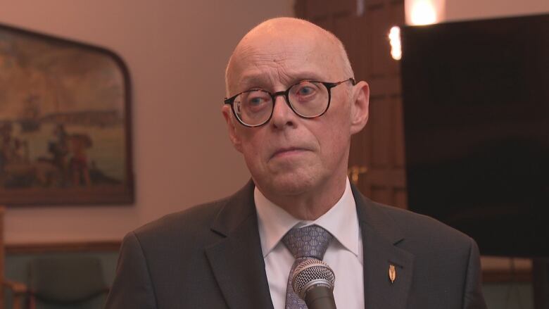 An older man wearing glasses and a black suit stands in the media scrum area of Confederation Building.