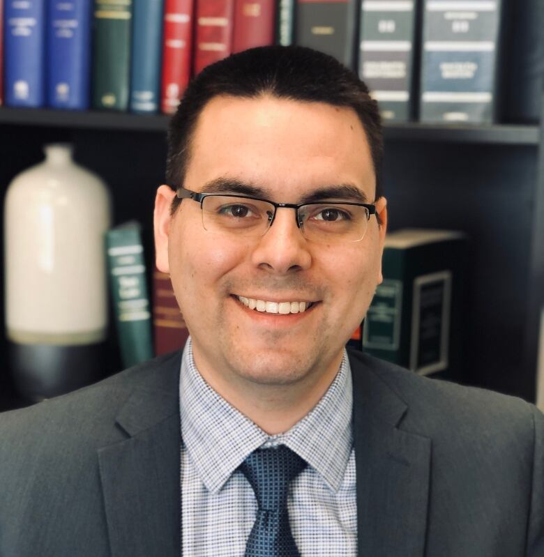 A dark-haired man wearing glasses is smiling. He is standing in front of a bookcase. 