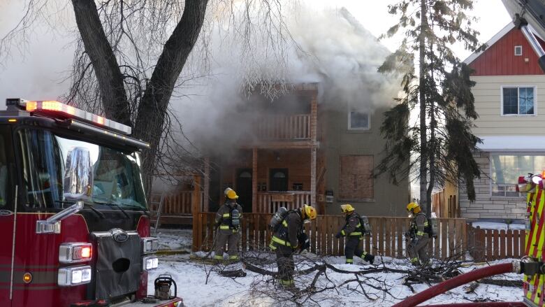 Four firefighters stand in front of a home covered by smoke. A red firetruck is in front of the home. 