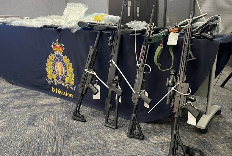 Long assault rifles lean against a table that has bags of drugs on it.