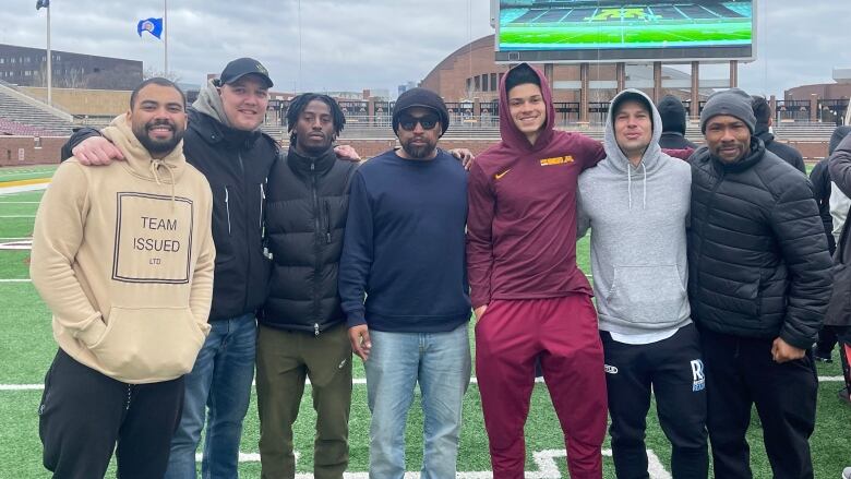 Seven people stand in a line looking at the camera on a football field.