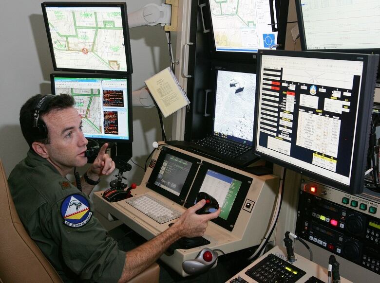 A man sits in front of numerous computer monitors illuminated with displays. He points with his left-hand and holds a control stick with his right.