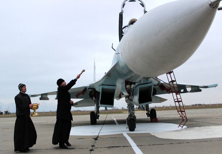 Two men in dark, long clothes stand near a military jet on a runway. One holds a chalice while the other nearer the aircraft extends a holy water sprinkler, or aspergillum.