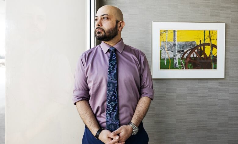 A man with a beard wearing a blue tie and purple shirt stands by a window. He looks off to the side with a solemn expression on his face.