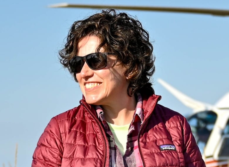 A woman with dark curly hair, sunglasses, and a red jacket smiles.