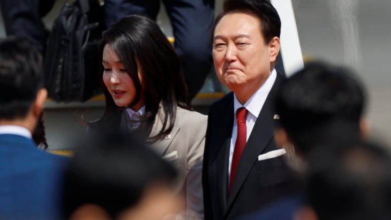 A man in a blue suit and red tie disembarks from an airplane. To his right is a woman wearing a grey blazer.