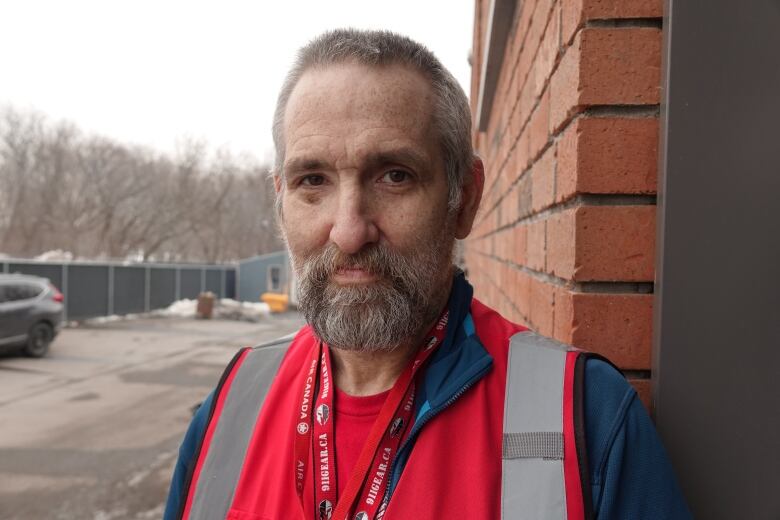 A man with short grey hair and a trimmed brown and grey beard stares at the camera. He as a black eye that's healing and is wearing a red vest and lanyards holding his ID cards.