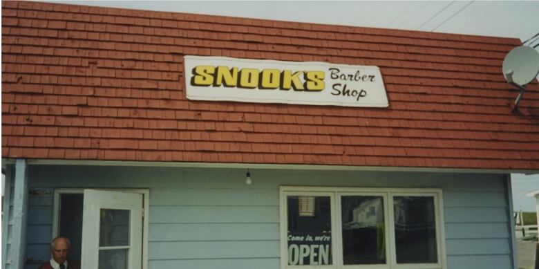 A photo of a barber shop. The building is blue with a red roof, and it's owner is standing in the doorframe.