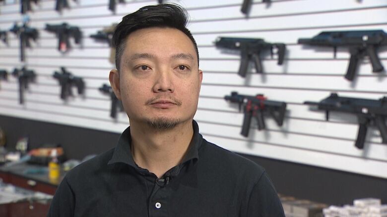 a man in a black polo shirt stands in front of a row of airsoft guns 