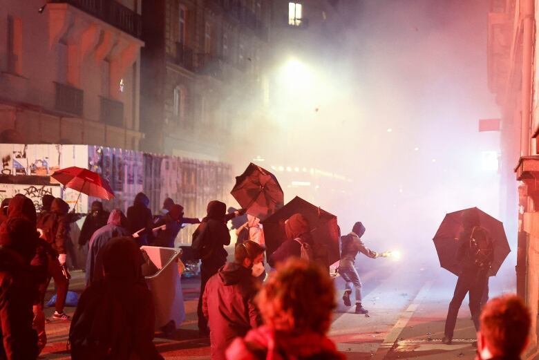 People holding umbrellas up against projectiles are lit up against a cloud of tear gas.