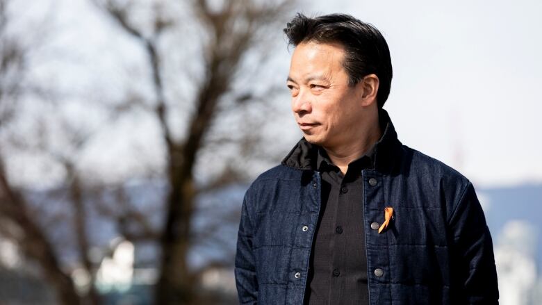 Vancouver's Mayor Ken Sim is pictured looking in the distance, to his right. An orange ribbon is pinned near his collar.