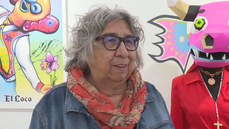 An Indigenous woman with gray hair and glasses stands wearing a red scarf and a denim shirt.