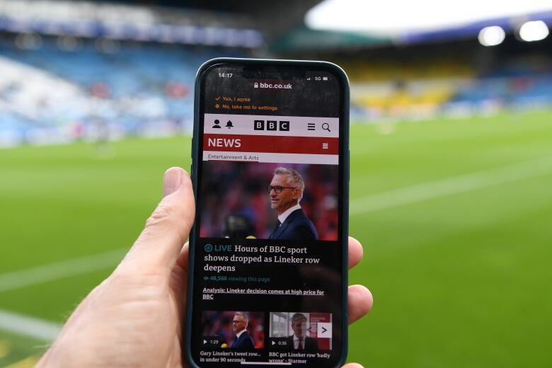 A hand holds up a cellphone in front of a soccer pitch. The screen shows a news website, with the main headline reading: LIVE: Hours of BBC sport hours dropped as Lineker row deepens.