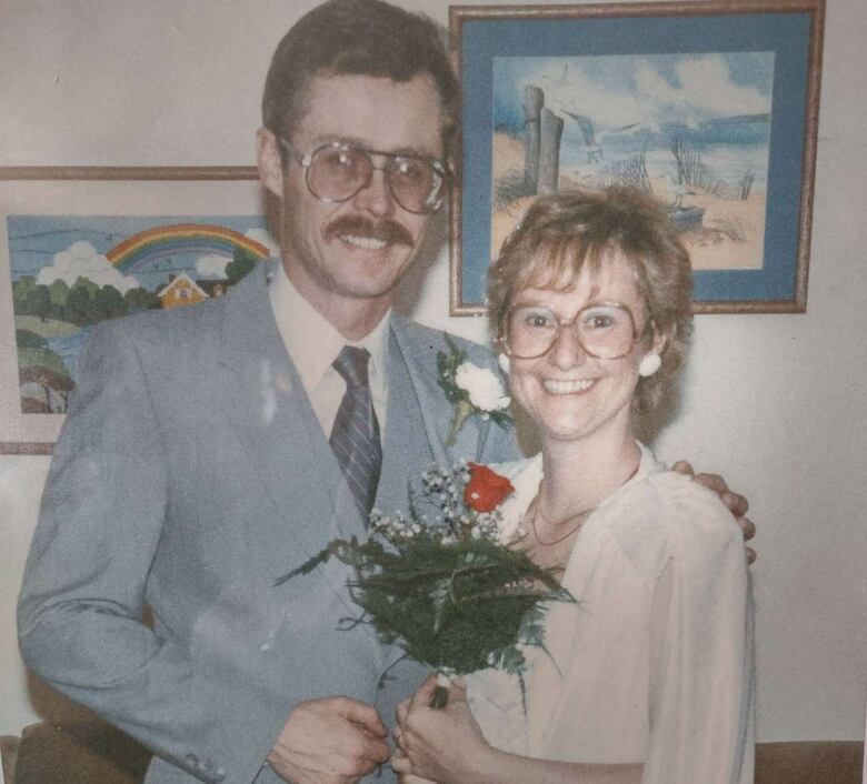 A man in a grey suit and a woman in a pink dress smile into the camera, while she holds a red rose.