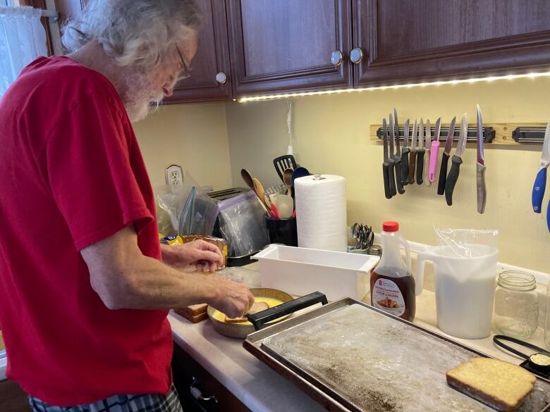 An older man in a red T-shirt makes french toast on a griddle.