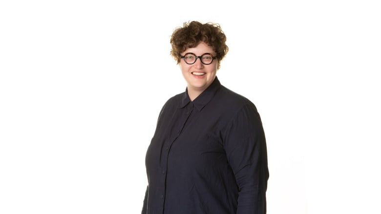 A woman with short curly hair and black-rimmed eyeglasses is smiling and looking directly at the camera, wearing a black collared shirt and standing against a white background. 